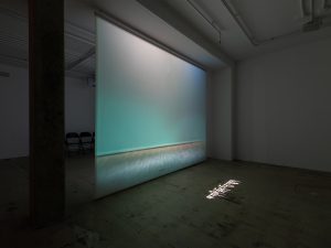 same room as the one above, a line of chairs sit in the back of the room, the projected image is green here, with a line of Tibetan text on the floor