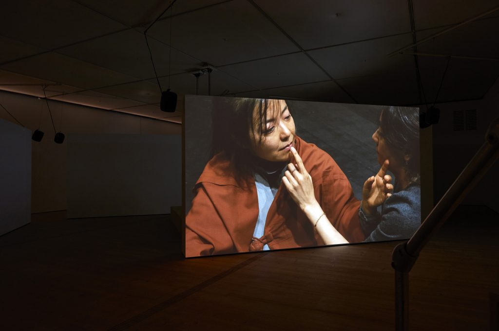 three of the four projection screens that stand alone on the floor are visible here, with one showing image of two East Asian Women engaged in showing affection towards each other sitting on the stage