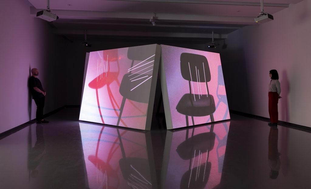 two wall-like surfaces used for projecting the images of the work lean slightly onto each other in a pink lit room, two people stands on opposite sides of the four channel video installation