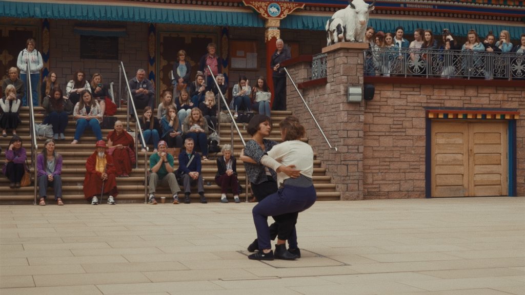 a close up of two female performers are engaged in an embraced position in front of a Tibetan Buddhist temple in Scotland, on its stairs sit a group of audience consists of the abbot and local visitors