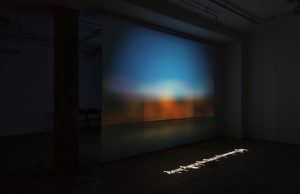 a darkened room, the projected image is suspended in the middle, the lighting on the image is blue on top with redish brown at the bottom, a line of Tibetan text is projected on the floor.