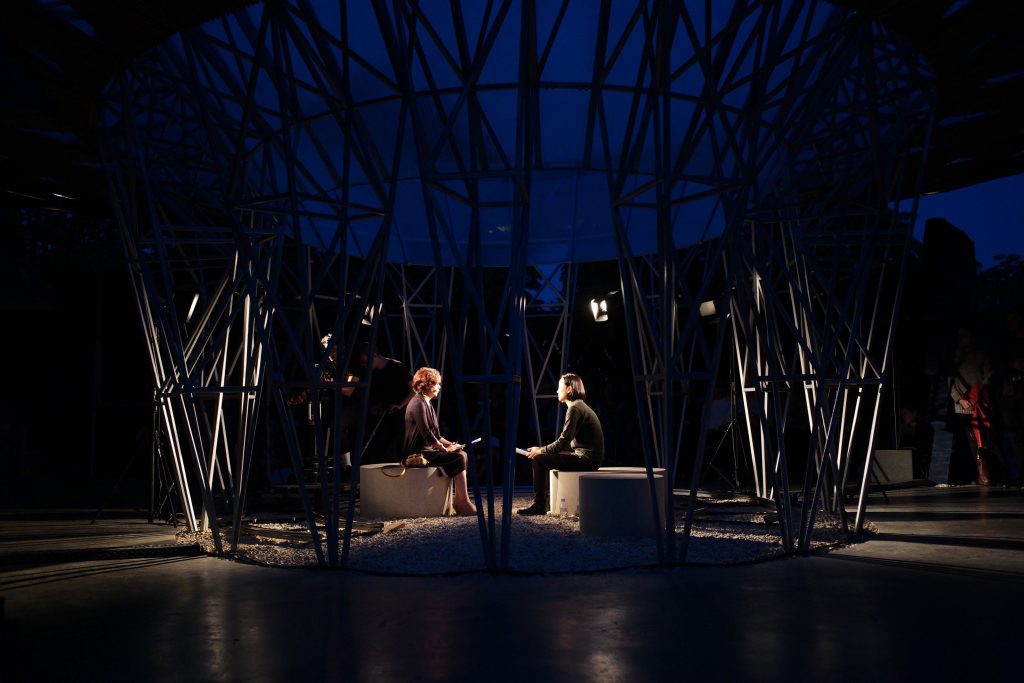 Shen Xin and Sungmee Kim sitting in the middle of the Serpentine Pavilion designed by Francis Kéré, engaged in a performance of conversations and translations, the sky is dark, the centre of the performance is lit