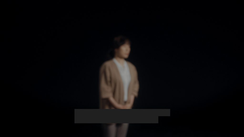 a blurred image of an East Asian woman wearing light brown jacket and white shirt standing against a dark background