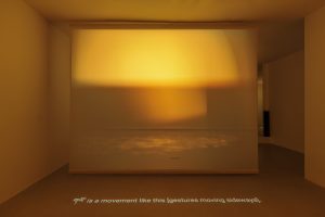 Contents: "a front facing image of the installation's yellow and orange phase, swinging theatre light on stage in the image, with a line of text projected on the floor that explains a Tibetan word as a movement.