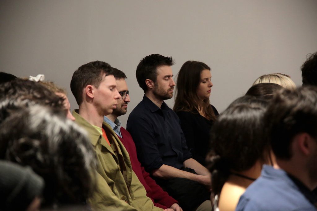 the camera here is focused on a few people in a crowd of seated people, their eyes are closed, following the instruction of a meditation.