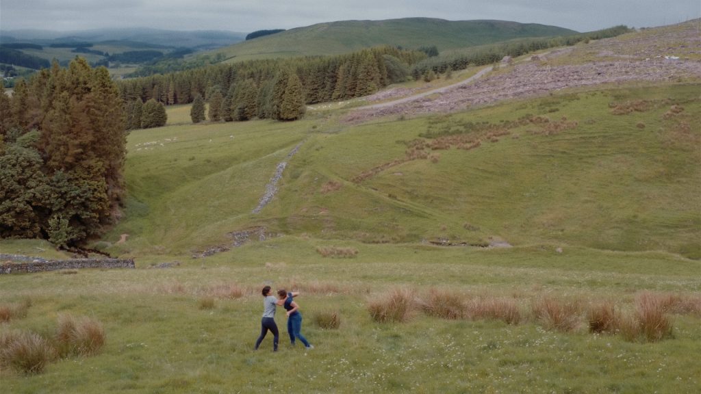 two female performers wearing t-shirts and jeans are engaged in a fighting position standing against fields of green grass and trees