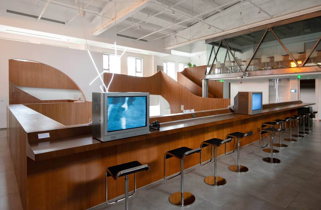 Two square monitors sit on a long brown table surface with brown high stools, agains an architecture space outlined by curves and circles