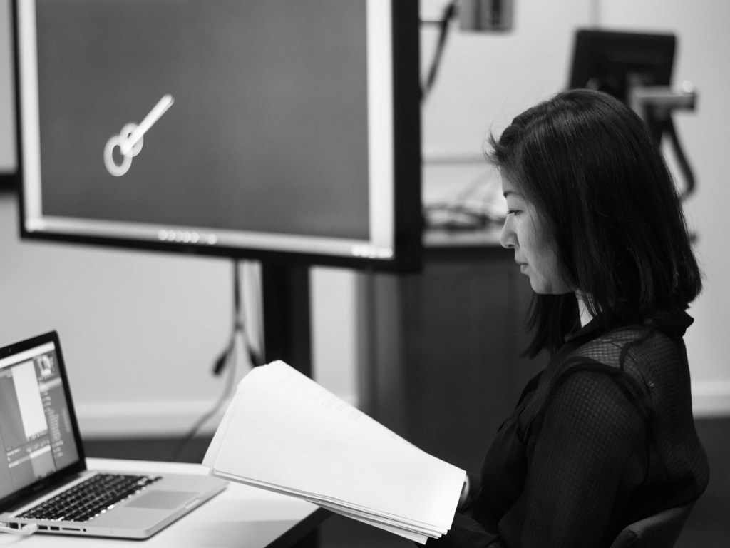 an asian woman holding a script facing a laptop connected to a tv screen showing an animation